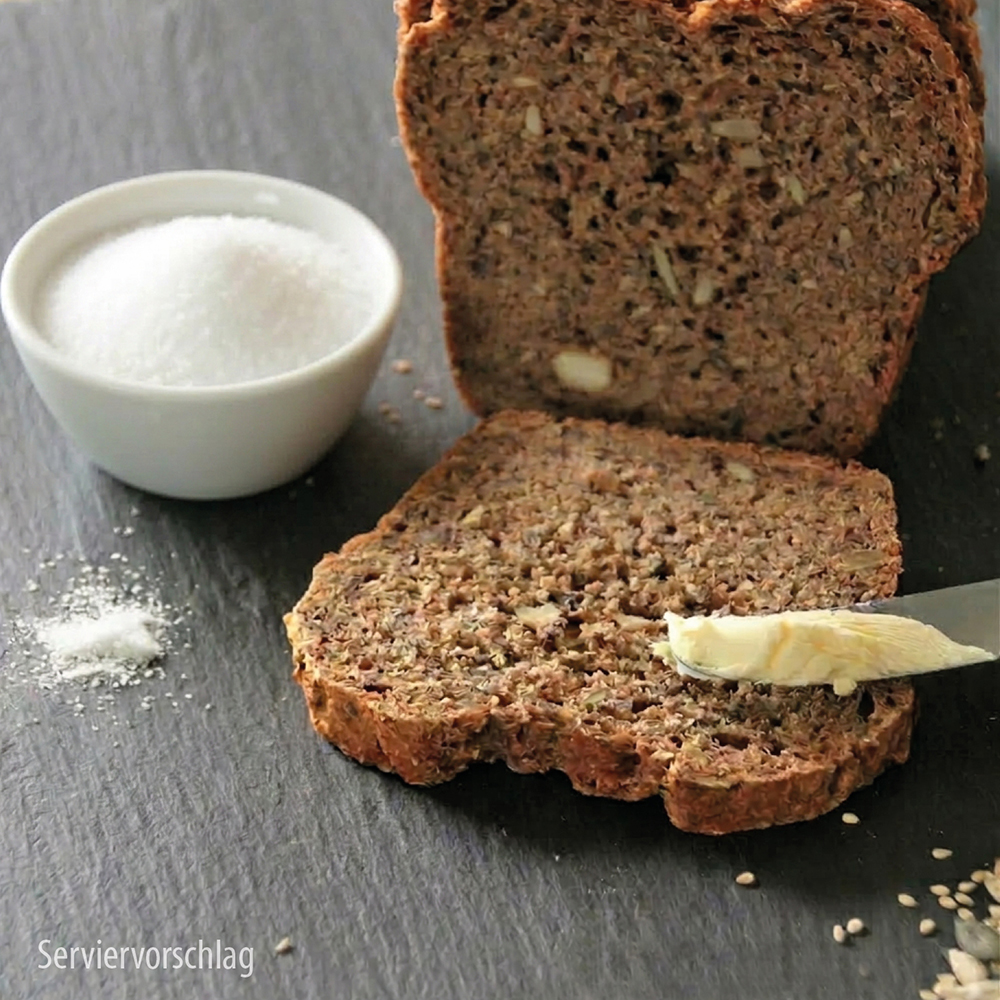 Eine Scheibe dunkles Vollkornbrot mit Kernen auf einer dunklen Schieferoberfläche, die mit einem Messer mit Butter bestrichen wird, dazu eine kleine Schüssel Salz und ein Laib Brot im Hintergrund. Serviervorschlag für Dr. Jacob’s Produkte.